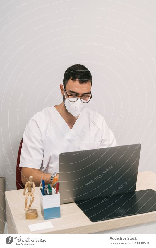 Serious doctor in medical room in hospital man work laptop mask clinic typing physician male busy occupation specialist device using table gadget modern