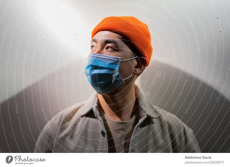 Serious Asian man in face mask standing in gray corridor respirator concentrate thoughtful pensive calm cover mouth coronavirus covid 19 ponder focus room