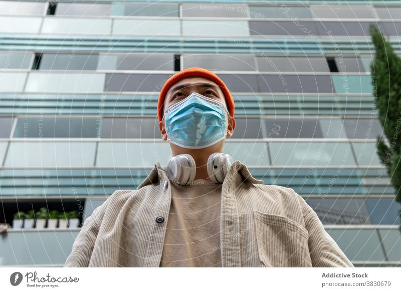 Emotionless Asian man in face mask standing against modern building emotionless urban condominium cover mouth headphones respirator coronavirus facade