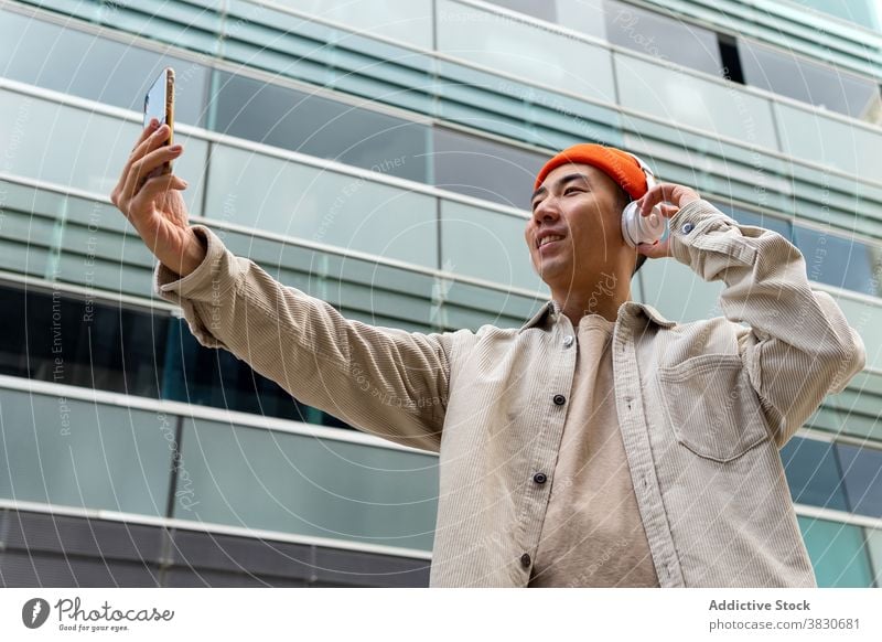 Joyful Asian man having video call outside modern building smartphone conversation talk headphones chat joy video chat happy condominium wireless casual facade