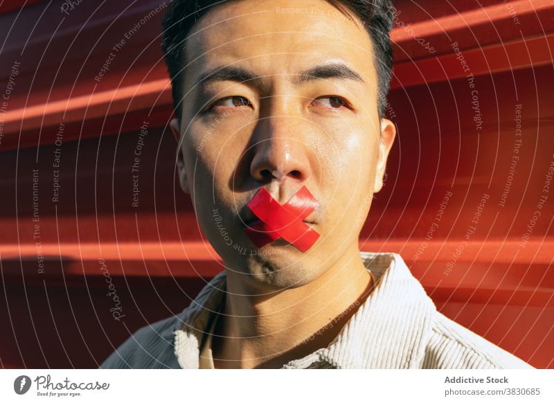 Crop Asian man with adhesive tape on lips hush seal muteness concept silent contemplate thoughtful voiceless pensive concentrate serious quiet stand plaster