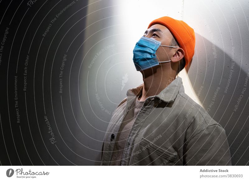 Serious Asian man in face mask standing in gray corridor respirator concentrate thoughtful pensive calm cover mouth coronavirus covid 19 ponder focus room