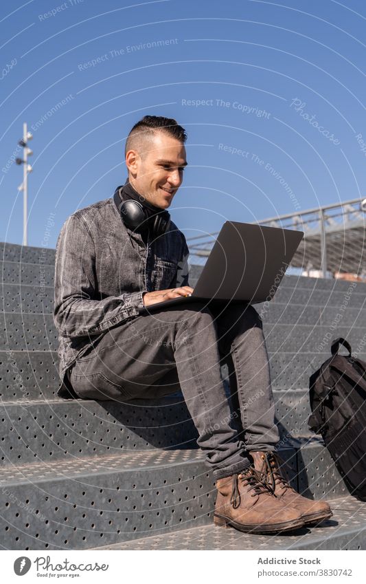 Cheerful young man working on laptop in headphones using typing cheerful internet freelance browsing stairs occupation male remote device modern surfing