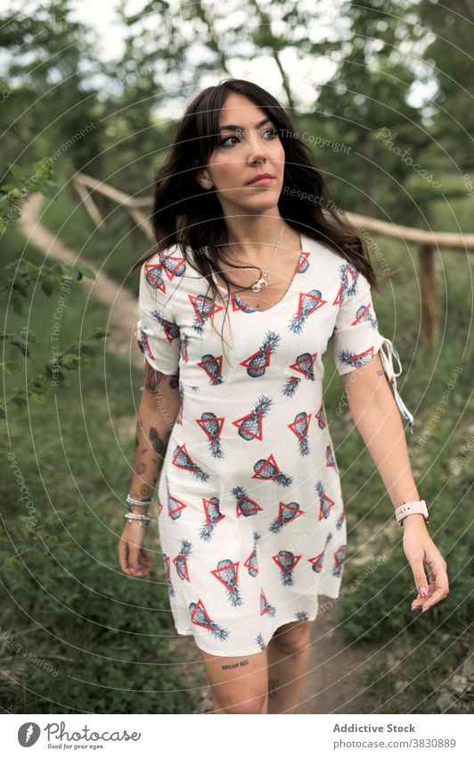 Dreamy woman walking on footpath along fence thoughtful stroll forest park dreamy style fashion nature female dress tattoo pensive serious dark hair accessory