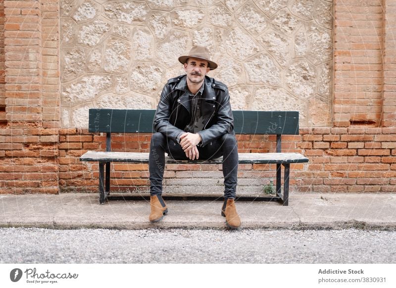 Cool hipster guy resting on bench man trendy dandy serious mustache hat urban leather jacket male brick wall stone hands together street city calm metrosexual