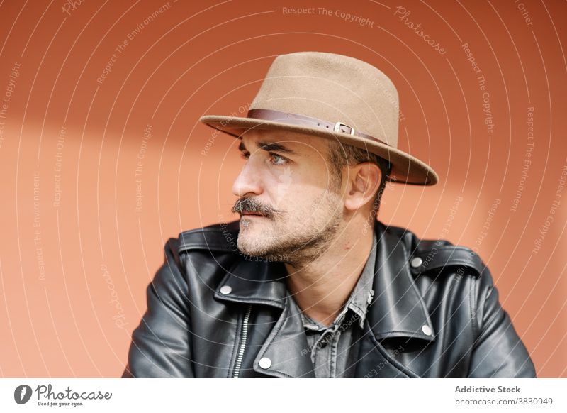 Pensive hipster with mustache standing on colorful wall man cool trendy leather jacket metrosexual attentive male hat arrogant thoughtful confident outfit
