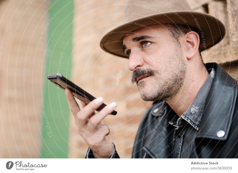 Pensive thoughtful trendy man recording message on smartphone mustache voice mobile beard hat leather jacket male building using dandy handsome style talk tell