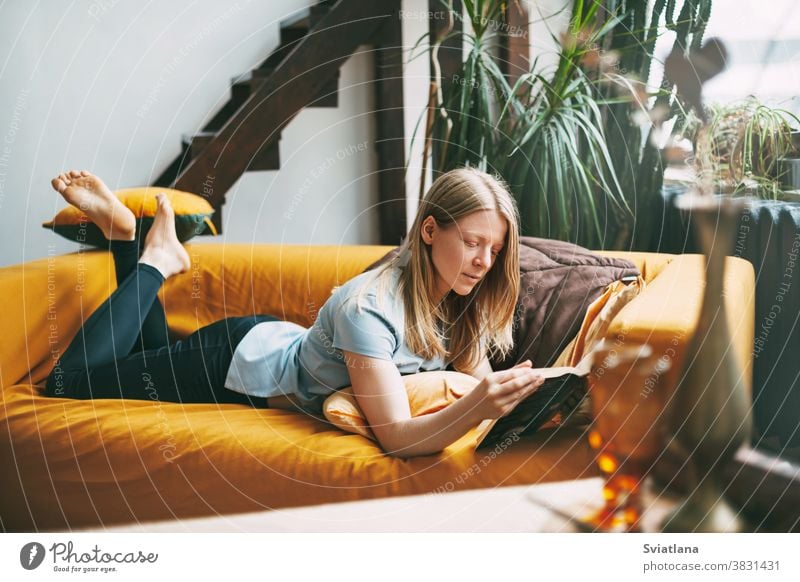 A young woman is lying on the sofa and reading a favorite book. The girl is resting, devotes her free time to reading. happy living novel indoors leisure home
