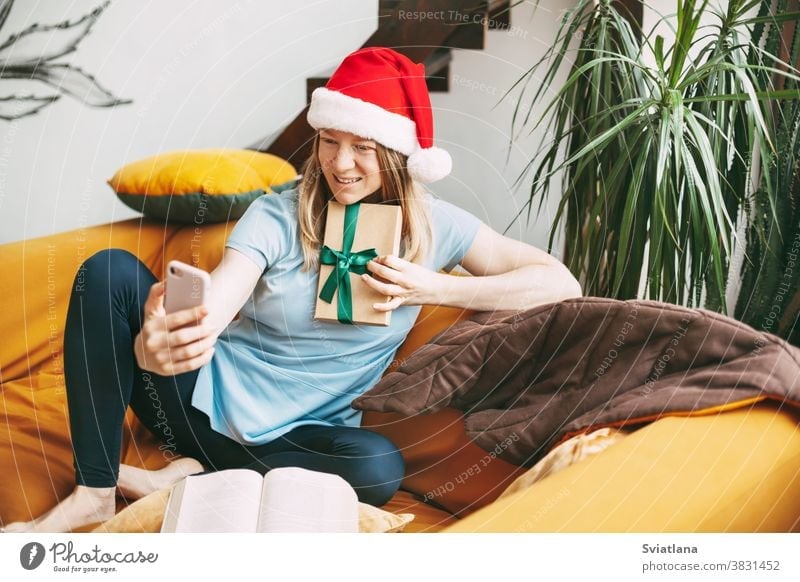 A smiling girl in a Christmas hat sits on the sofa and communicates via video link with friends and shows them her gift. Girl congratulates friends and family on holidays
