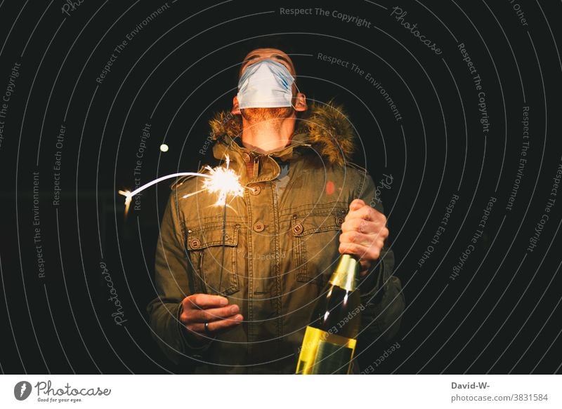 Man with a mouthguard / breathing mask stands outside on New Year's Eve and holds a sparkler and champagne bottle in his hands corona coronavirus