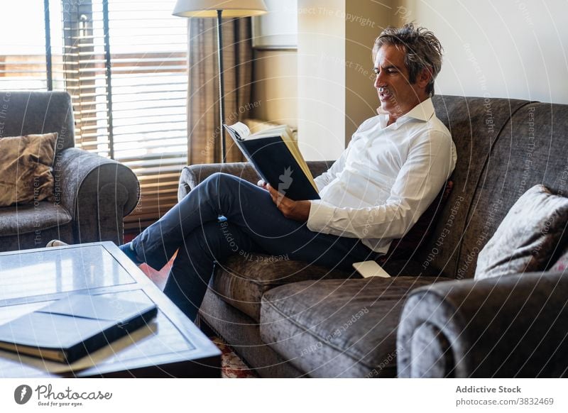 Mature man reading book at home sofa table sit male casual modern lifestyle focus mature adult relax rest smart story fiction concentrated novel literature