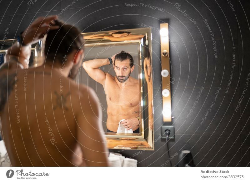 Shirtless man doing hair near mirror ponytail bathroom hairdo modern illuminate lamp shirtless reflection male adult beard tattoo routine home apartment hygiene