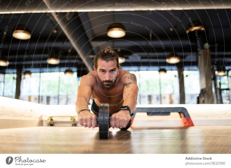 Bearded sportsman exercising with abs wheel exercise gym training floor modern shirtless strong male athlete adult physical strength effort wellness workout