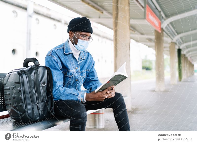 Ethnic man reading book on railway station wait train platform traveler mask coronavirus railroad male ethnic black african american trip new normal bench