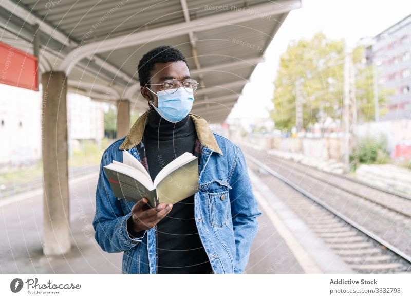 Black man with book on train platform wait railway new normal traveler coronavirus mask male ethnic black african american pandemic railroad transport station