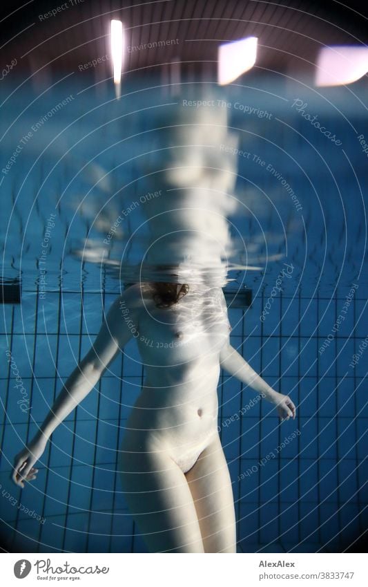 Underwater body portrait with reflection at the waterline of a young woman under water, standing naked in a pool and bathing Young woman pale underwater