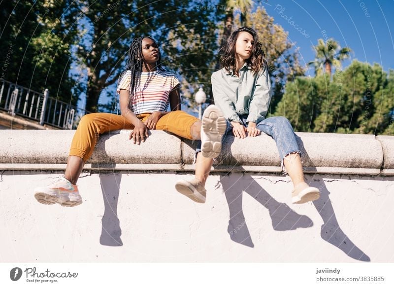 Two multiethnic women sitting on a urban wall. serious expression sunlight friend black afro girl student two people lifestyle female pretty young friendship