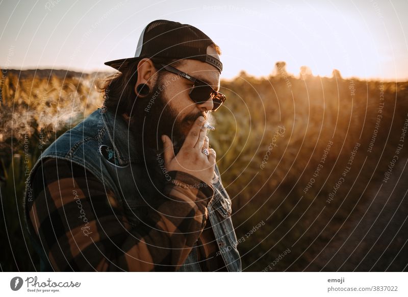 young man with beard, sunglasses and cap smokes teen Sunset out Field Hip & trendy Hipster Man Smoking Cigarette Sunglasses Headwear Unhealthy Lifestyle Cool