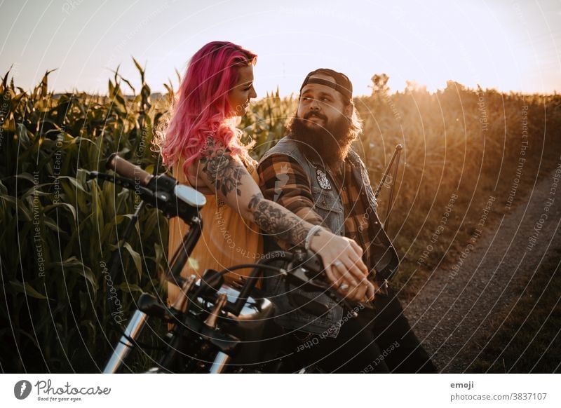 young couple with tattoos and pink hair and motorcycle teen Couple Love In love Sunset out Field Tattoo Hip & trendy Hipster Man Woman eye contact Maize field