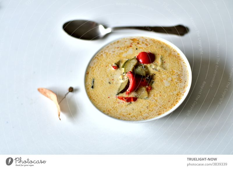 Non fat reduced coconut milk vegetable soup with red pepper, zucchini, cucumber and cherry tomato in a white soup bowl on a white table with a soup spoon and a dried lime blossom leaf