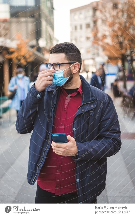 Man fixing the face mask while walking in the city, holding a smartphone adult communication coronavirus covid-19 epidemic european eyeglasses eyes freelancer