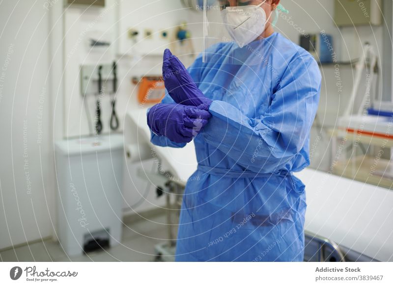 Anonymous doctor in uniform putting on sterile gloves put on job process equipment medical clinic woman mask respiratory hospital blue cap couch professional