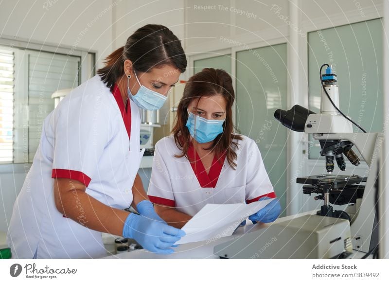 Anonymous medical technologists watching paper near microscope in lab scientist uniform focus colleague women partner professional clinic device staff hospital