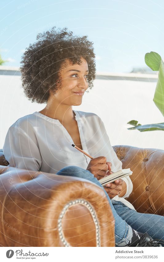 Happy ethnic woman taking notes in notepad idea write take note think memo terrace female black curly hair african american notebook armchair work contemplate