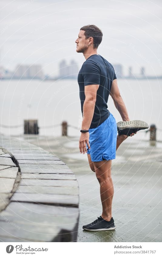 Male runner stretching legs during training warm up city man embankment fit sportsman wet street male forward bend exercise wellness flexible workout sportswear