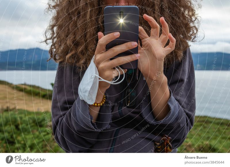 A young woman, grasping a face mask and a smart mobile phone, Spain. girl hair curly protective new normal chat looking internet digital telecommunications call