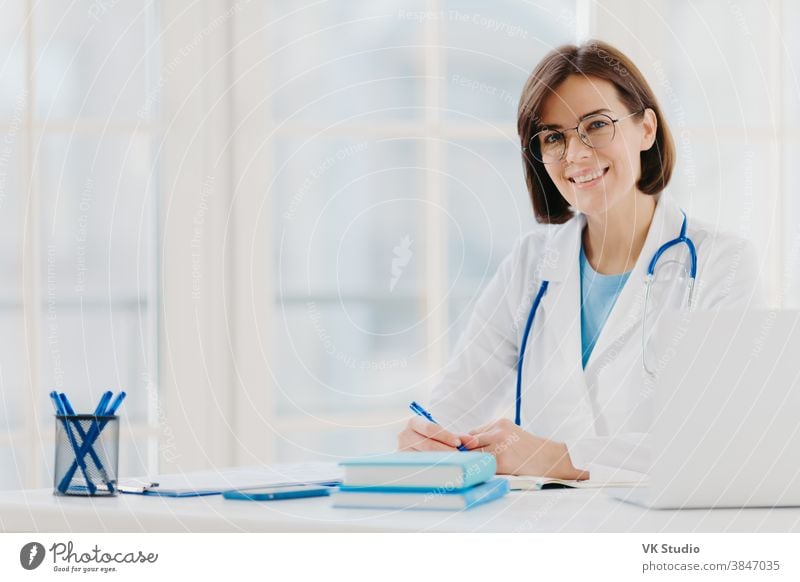 Professional woman doctor writes down notes, poses at desktop in office with laptop, wears white coat, spectacles and phonendoscope around neck, looks through medical documents. Healthcare concept