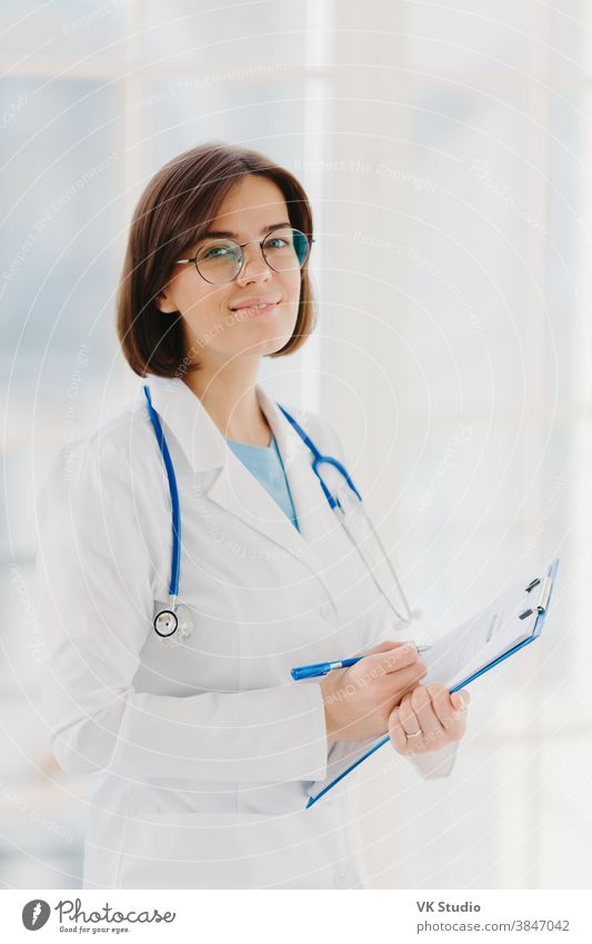 Qualified cardiac expert with stethoscope prepares for health seminar, gives consultancy about first aid, writes down information in clipboard, wears white gown, ready to give medic care help