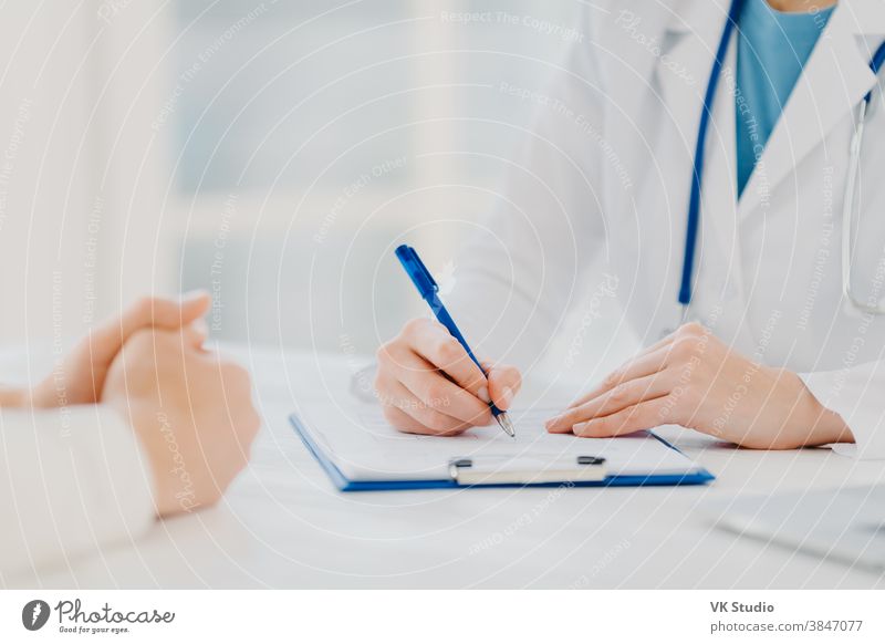 Unknown female doctor writes down prescription on papers in clipboard, consults patient about curing disease, pose at desktop against blurred white background fills up medical form. Symptoms diagnosis