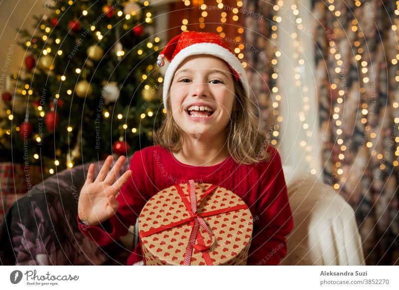 A happy child in red Santa hat is celebrating Christmas. Cute girl holding wrapped gift, smiling and waving hand, sitting in decorated with Christmas lights and tree room. Happy holidays