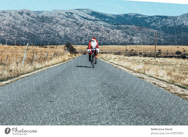 Santa Claus riding bicycle along mountain road santa claus ride bike empty costume red color male modern journey freedom speed trip move sunny activity
