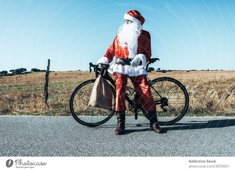 Santa Claus near bicycle on road in countryside santa claus bag relax roadway vehicle bike santa hat male beard costume red color modern style stand transport