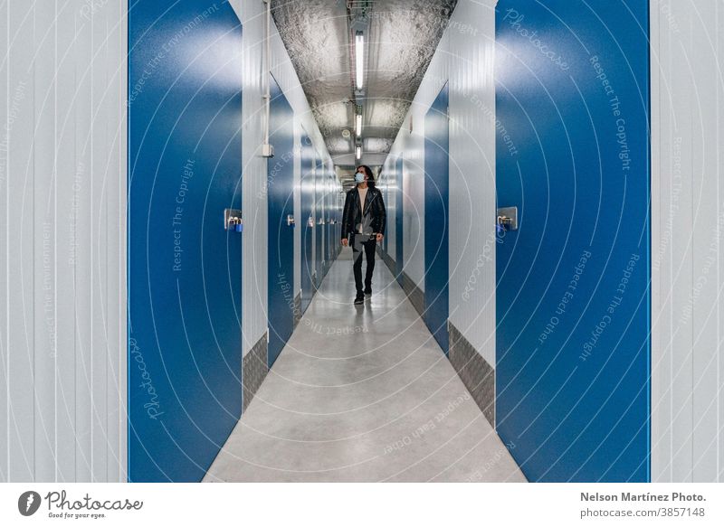 Hispanic man in a leather jacket walking in a hallway with blue doors and white walls. mask covid19 depht storage room lifestyle cool young work coronavirus