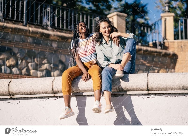 Two friends having fun together on the street sitting on a urban wall. women multiethnic black afro girl student two people lifestyle smile female pretty young