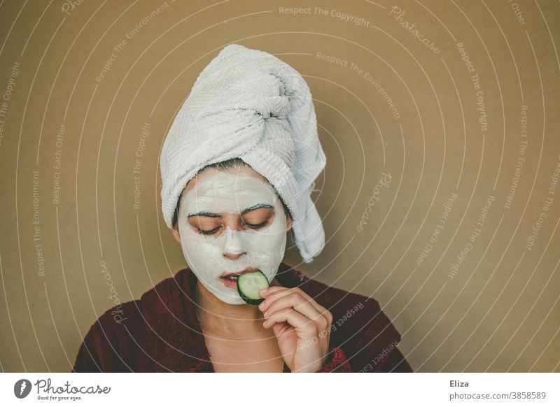 Woman with bathrobe and towel on her head enjoys a face mask and nibbles a slice of cucumber. Wellness, face care. Face mask cucumber slices Personal hygiene