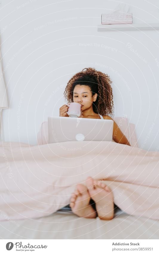Ethnic woman with cup of coffee and laptop sitting on bed bedroom drink bored at home relax rest free time young female african american black ethnic device