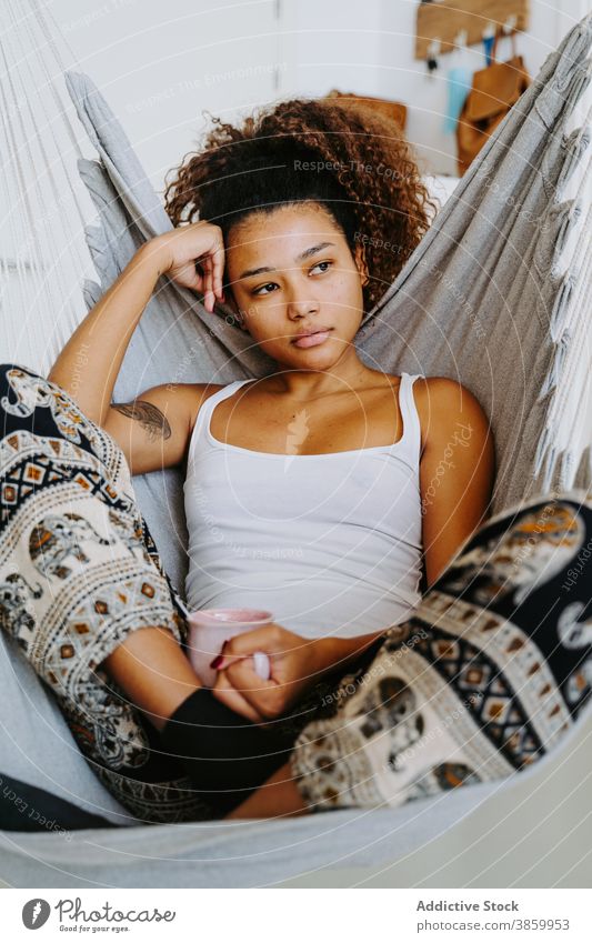 Ethnic woman with cup of coffee resting in hammock at home drink relax chill cozy young female african american black ethnic beverage lifestyle tea weekend