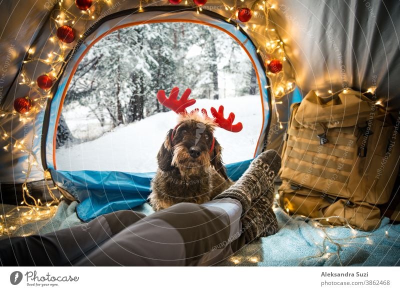 A man lying in a tent decorated with Christmas lights wearing warm wooden socks. Beautiful winter wild forest covered with snow. Self-isolation and social distancing during holidays. Travel with dog