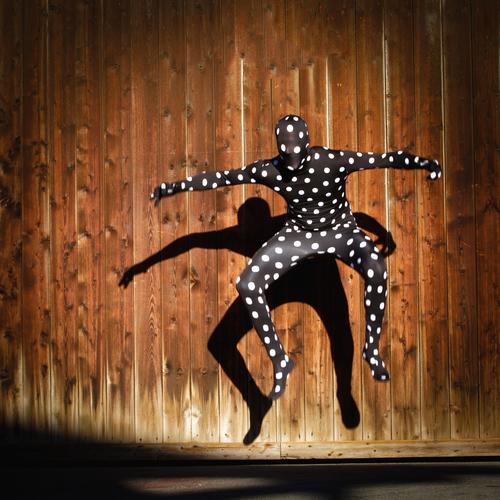 Human figure in dotted morphsuit costume, jumping into the air with shadow cast on a wooden wall Human being Morph Suite full body suit points in a dive