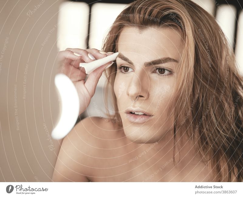 Androgynous male with long hair applying makeup man studio androgynous beauty foundation mirror transgender appearance young model facial pad visage routine