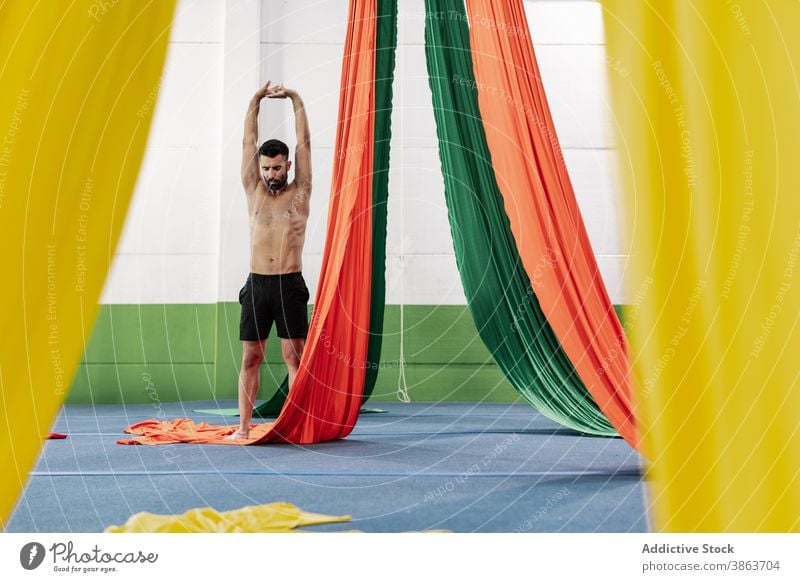 Shirtless male dancer stretching arms near aerial silks man rehearsal studio arms raised warm up practice flexible adult ribbon muscular perform colorful
