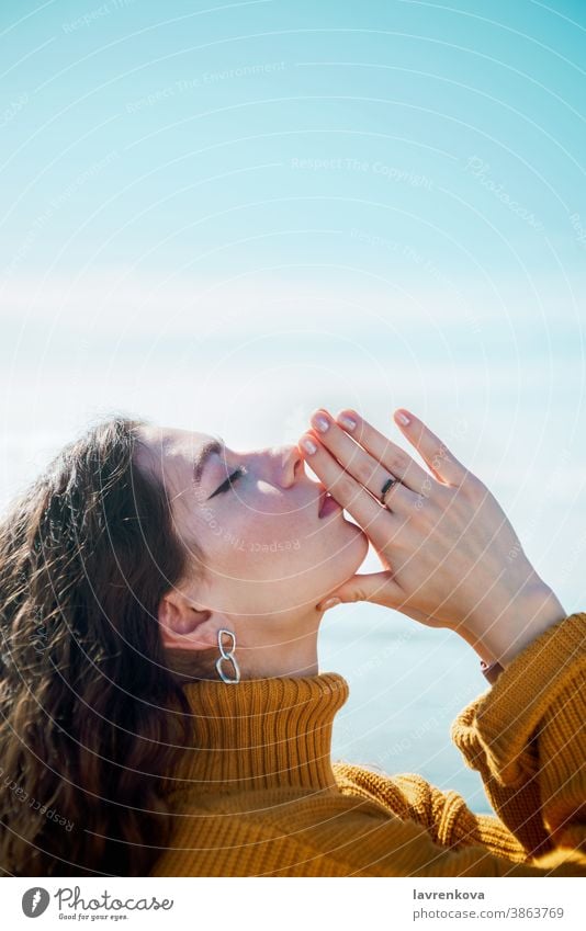 Profile portrait of young adult white female with eyes closed doing namaste gesture and touching her hands in front of blue skies relaxation practice lifestyle