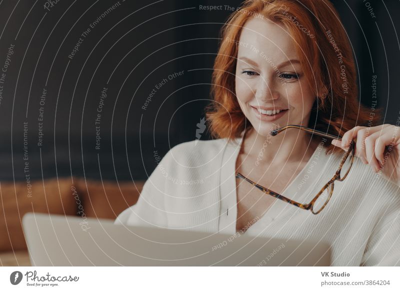 Beautiful redhaired woman concentrated in laptop display, smiles positively, holds optical glasses, poses indoor, uses modern technologies for online communication, watches webinar, reads news