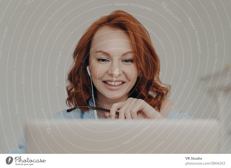 Pretty positive redhead woman gives online consultation, sits in front of opened laptop computer, has discussion with corporate client, smiles happily, uses modern earphones, enjoys online studying