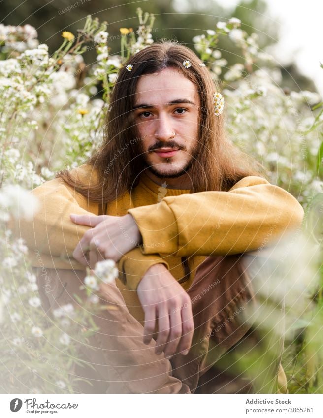 Young man with flowers in long hair sitting in field gay romantic bloom relax rest nature peaceful calm young male brunet lifestyle homosexual gender lgbt lgbtq