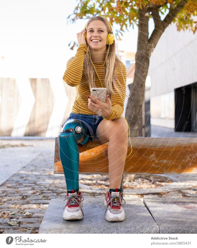 Cheerful woman with leg prosthesis listening to music in headphones bionic street city enjoy female song amputee sunny relax device happy cheerful smartphone